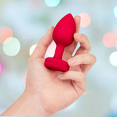 A hand holds the B-Vibe Vibrating Heart Plug Medium/Large - Scarlet Ruby against a softly blurred, multicolored background with circular light spots, perfect for exploring anal play.